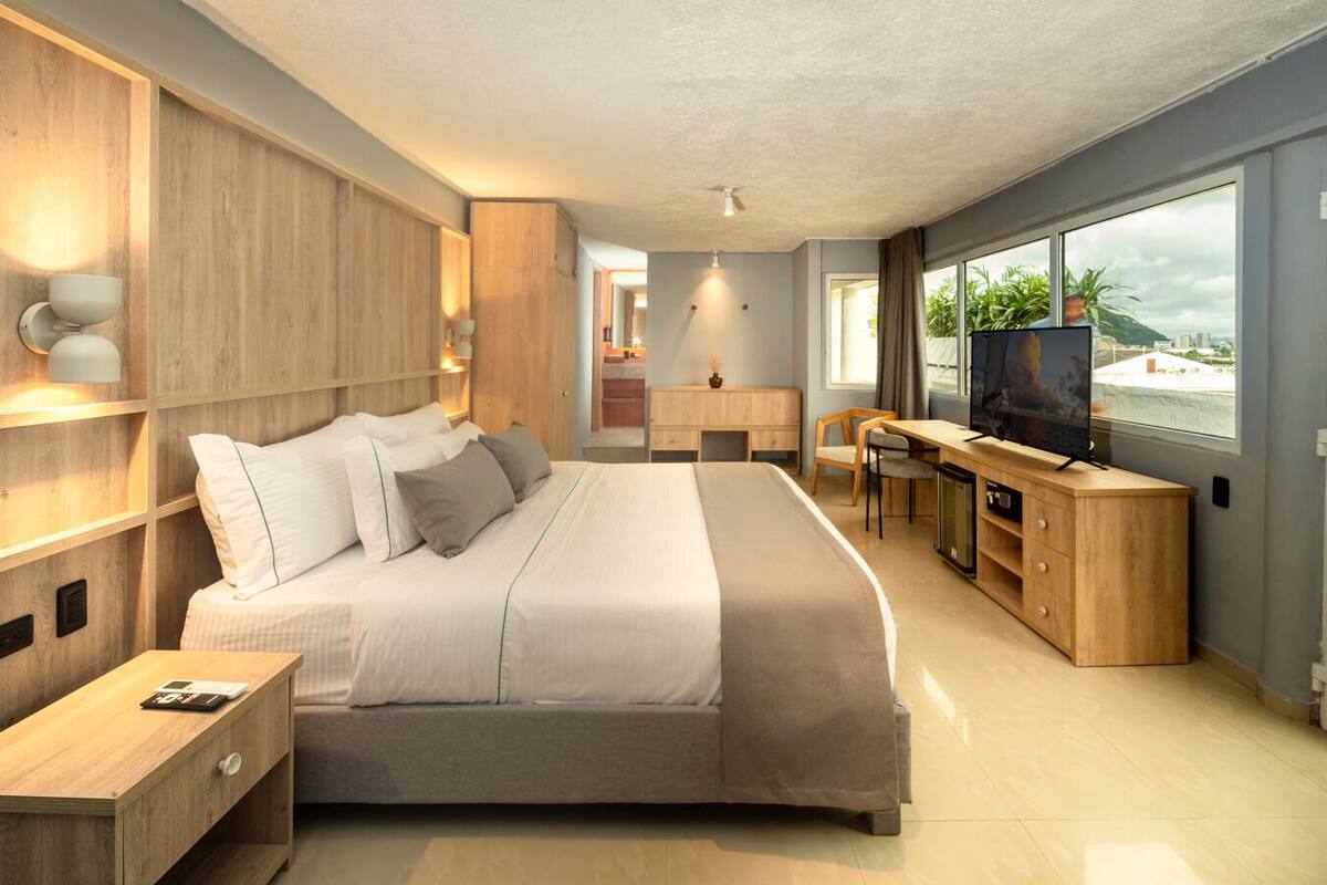 Quarto moderno com cama grande, mesas de cabeceira, escrivaninha e televisão.