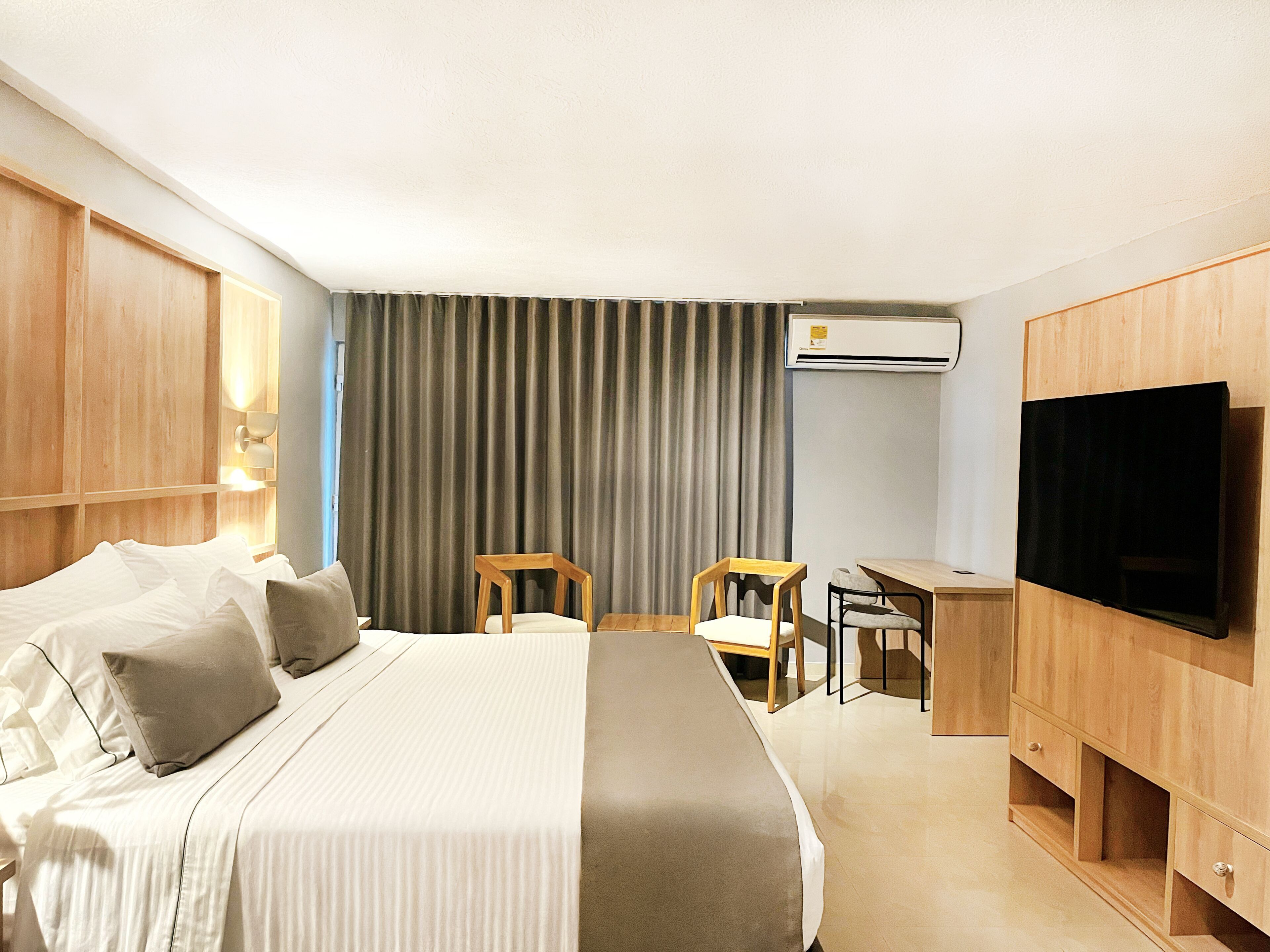 A hotel room with a bed, a desk, a chair, a television, and an air conditioning unit.