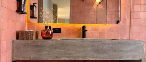 A bathroom with a large mirror, a wooden vanity, a vase with dried flowers, and a wall-mounted light fixture.