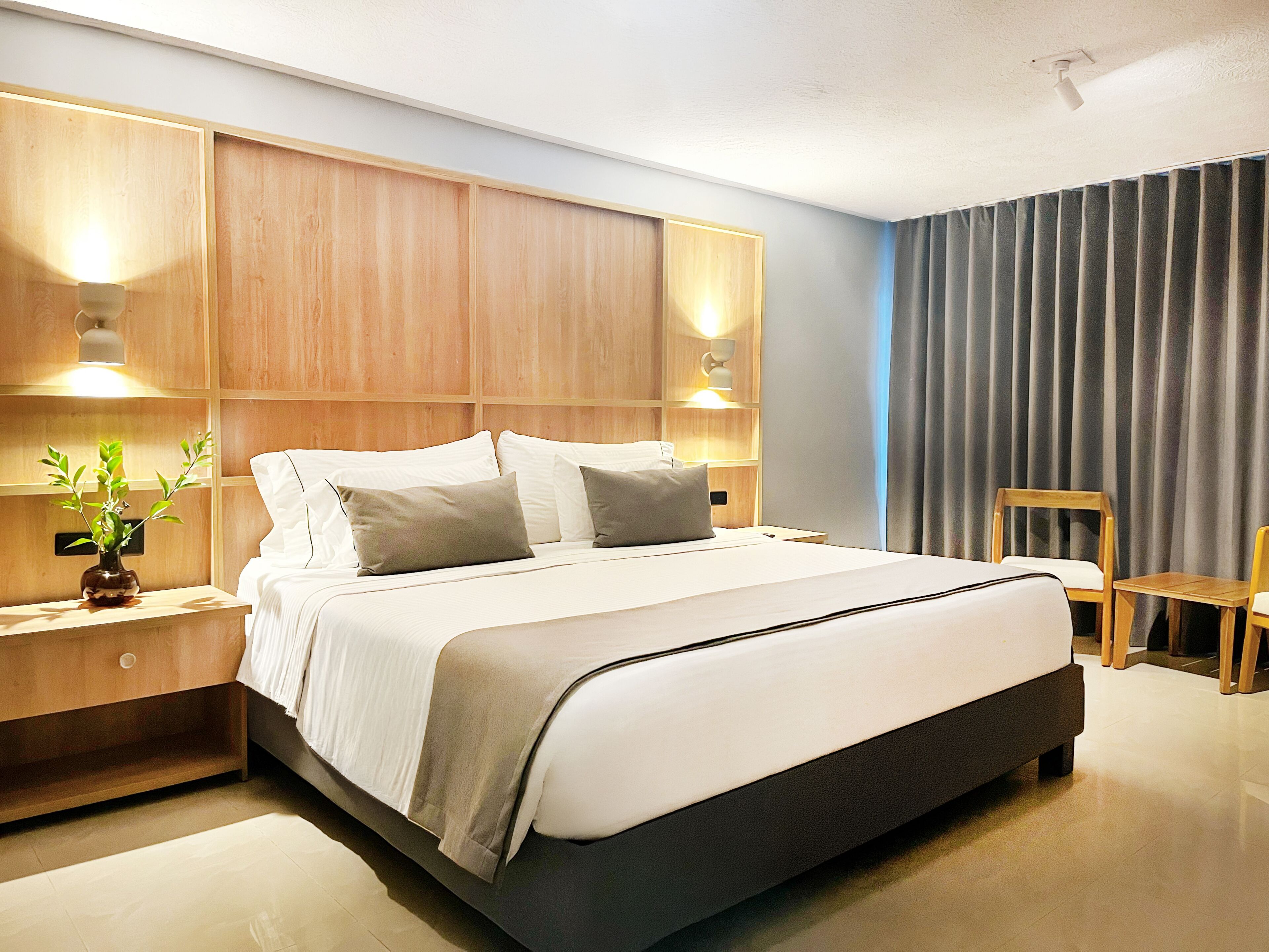 Une chambre d’hôtel moderne avec un grand lit, une tête de lit en bois, des tables de chevet, une petite table de nuit avec une plante et une chaise.