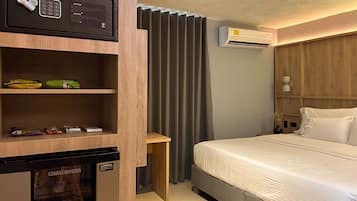 Una habitación de hotel con cama, un minibar, una mesita, una cortina y un equipo de aire acondicionado.