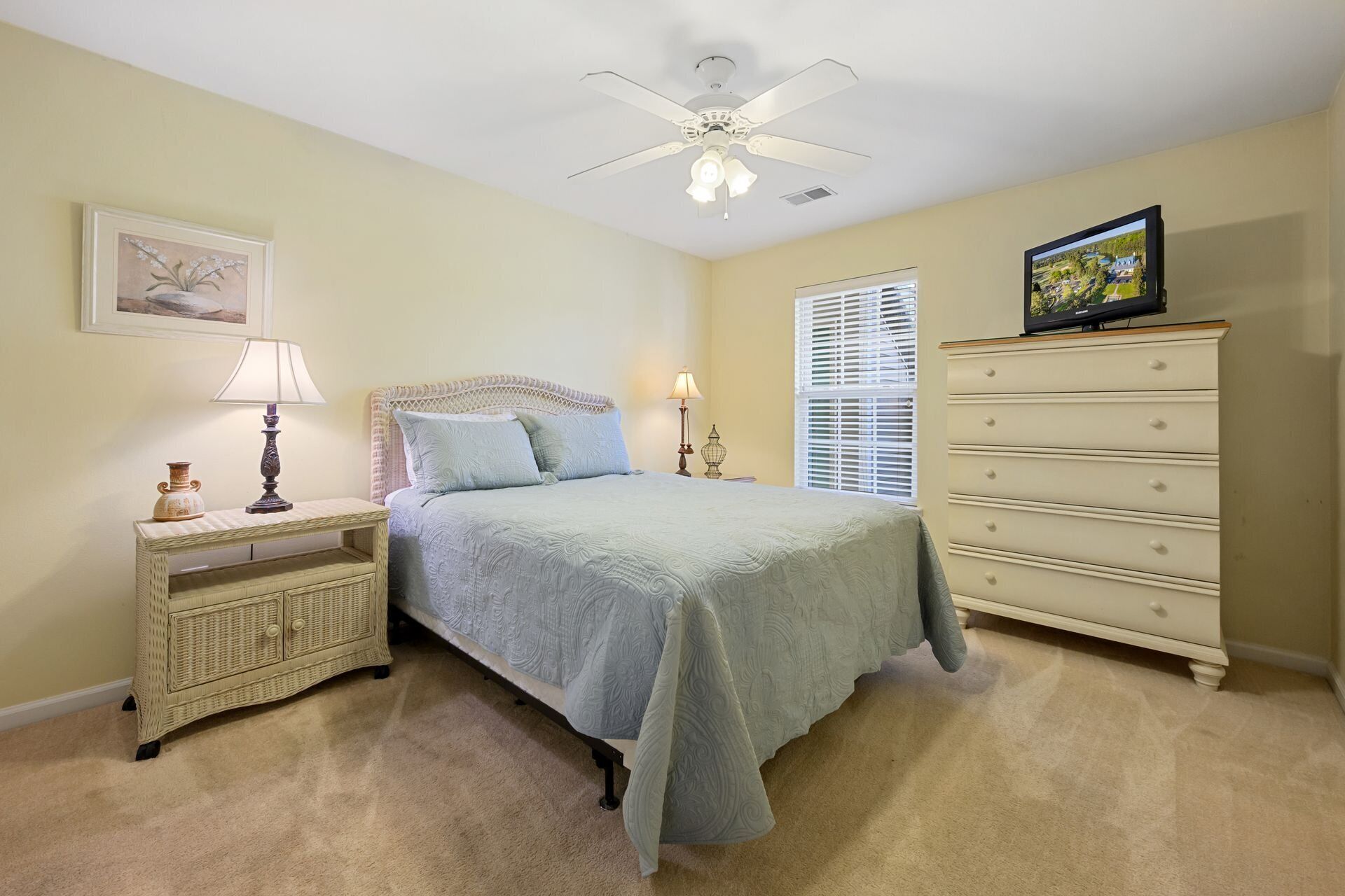 Une chambre à coucher comprenant un lit, une table de chevet, une commode, un ventilateur de plafond, un téléviseur et une fenêtre avec des stores.