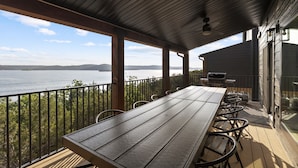 A covered outdoor deck with a long table and chairs, overlooking a body of water and distant mountains. - Lakefront | Three homes (sleeps 68) golf simulators, outdoor saunas, & hot tubs (HOLLISTER)