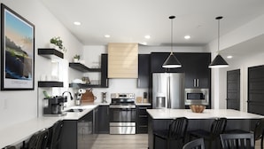 A modern kitchen with a central island, stainless steel appliances, black cabinetry, and pendant lighting. - Lakefront | Three homes (sleeps 68) golf simulators, outdoor saunas, & hot tubs (HOLLISTER)
