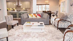 A spacious living room with a grey sofa, patterned armchairs, and a white coffee table. A kitchen is visible in the background with modern cabinetry and pendant lighting. - Modern comfort within walking distance from UKY, Kroger Field, & Chevy Chase. (Lexington)