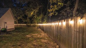 A wooden fence with string lights, a grassy yard, and a building with a sloped roof. - Modern comfort within walking distance from UKY, Kroger Field, & Chevy Chase. (Lexington)