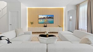 Una sala de estar moderna con un televisor de pantalla plana grande montado en una pared de paneles de madera, un conjunto de sofá blanco, una mesa de centro con un jarrón de flores y una escalera a la izquierda.