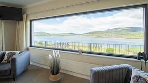 A room with a large window overlooking a lake and hills, a sofa, and a potted plant. - Dingle Bay Nest Burnham (Ventry)