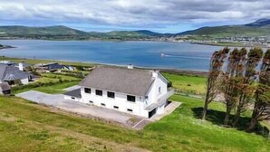 A white house with a grey roof, situated on a grassy hill overlooking a large body of water and distant hills. - Dingle Bay Nest Burnham (Ventry)