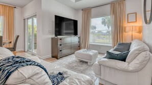 A modern living room with a flat-screen TV, a white sofa, a white armchair, and a large window with curtains.