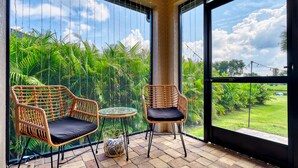 A balcony with wicker chairs and a small table, overlooking a lush garden and a clear sky.