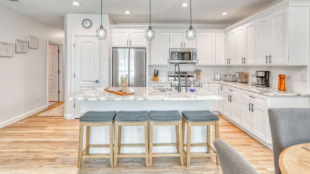 A modern kitchen with white cabinets, a stainless steel refrigerator, a microwave, and a sink. There are three bar stools and a dining table.
