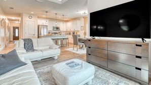 A modern living room with a flat-screen TV, a sofa, a coffee table, and a dining area with a kitchen in the background.