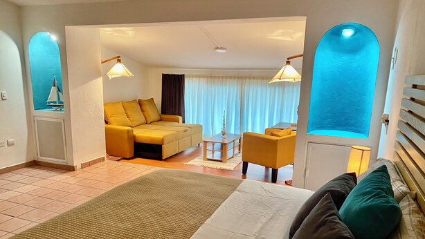 A hotel room with a bed, a sofa, a dining table, and a chair. There is a window with curtains and a view of the outside. - Come In (Puerto Vallarta)