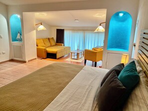 Ein Hotelzimmer mit Bett, Sofa, Esstisch und Stuhl. Ein Fenster mit Vorhängen bietet einen Blick nach draußen.