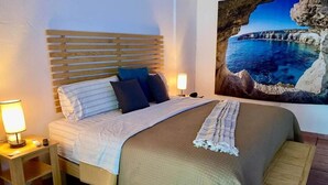A neatly made bed with a headboard, two bedside tables with lamps, a blanket, and a painting of a coastal scene. - Come In (Puerto Vallarta)