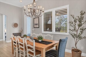 A dining room with a wooden table, white chairs, a chandelier, and a window with blinds.