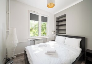 A modern bedroom with a large bed, a window with blinds, a radiator, and a wall-mounted shelf.