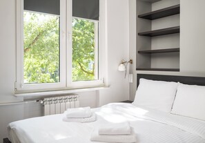 A neatly made bed with white linens and towels, a window with a view of trees, and a wall-mounted shelf with a small plant. - Nowa Praga Comfort Apartment (Warszawa)