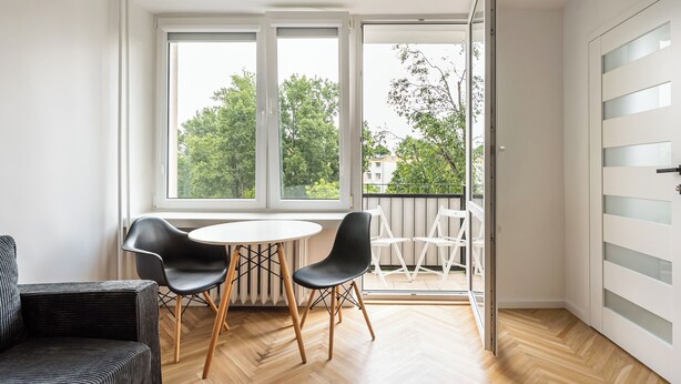 A modern room with a round table and two chairs, large windows with a view of trees and buildings, and a sliding glass door leading to an outdoor balcony. - Nowa Praga Standard Apartment (Warszawa)