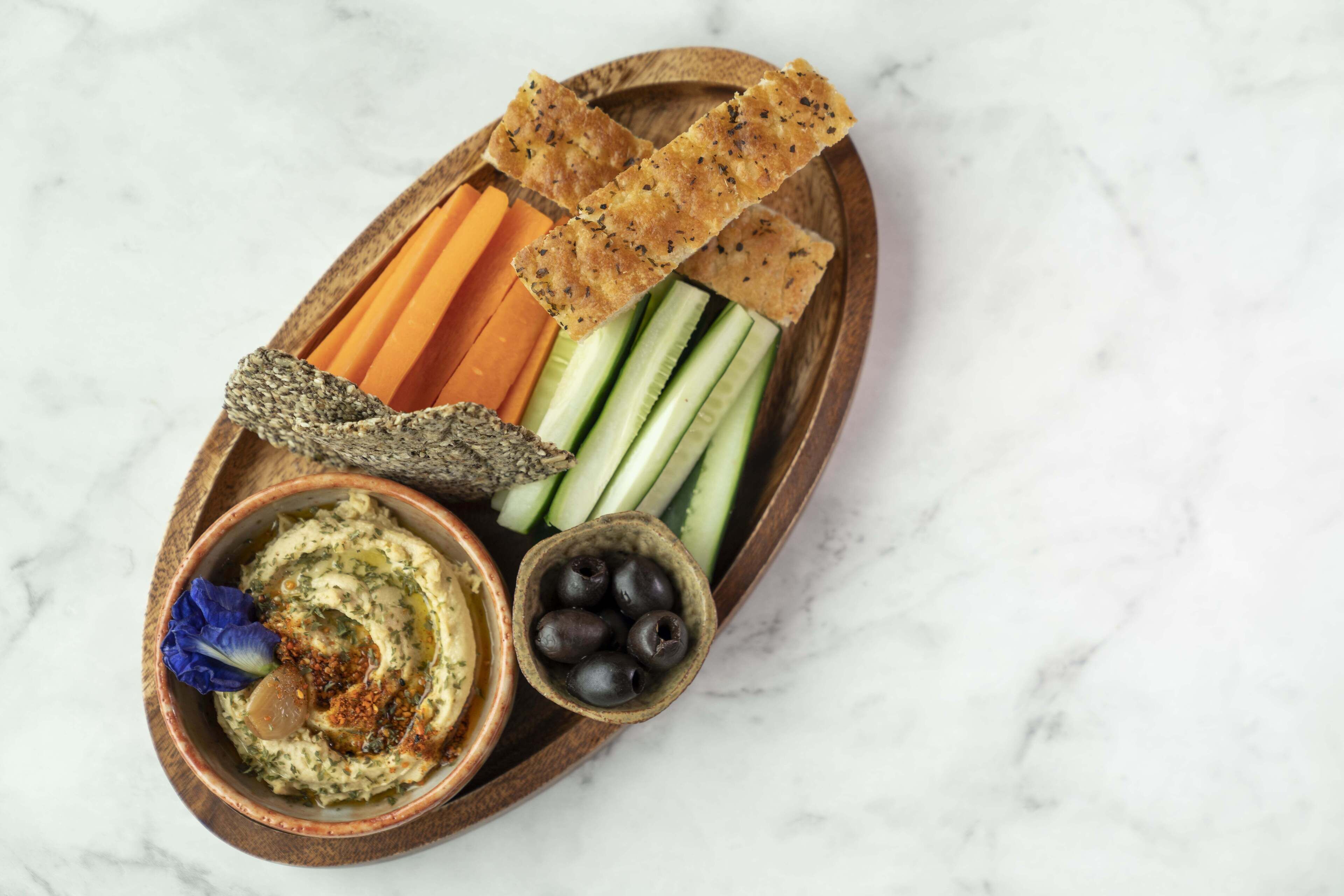 Un plateau en bois garni de houmous, d’olives, de carottes, de concombres et de crackers.
