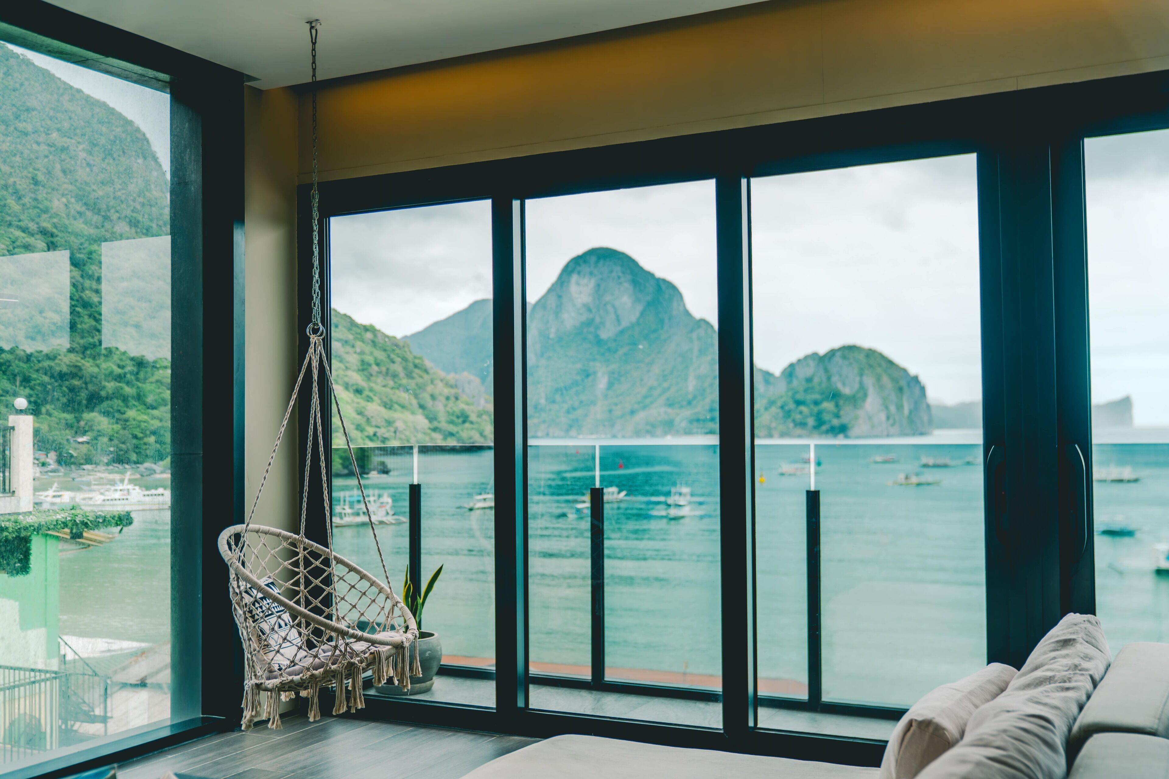 A room with a large window overlooking a body of water and mountains, a hanging chair, and a sofa.