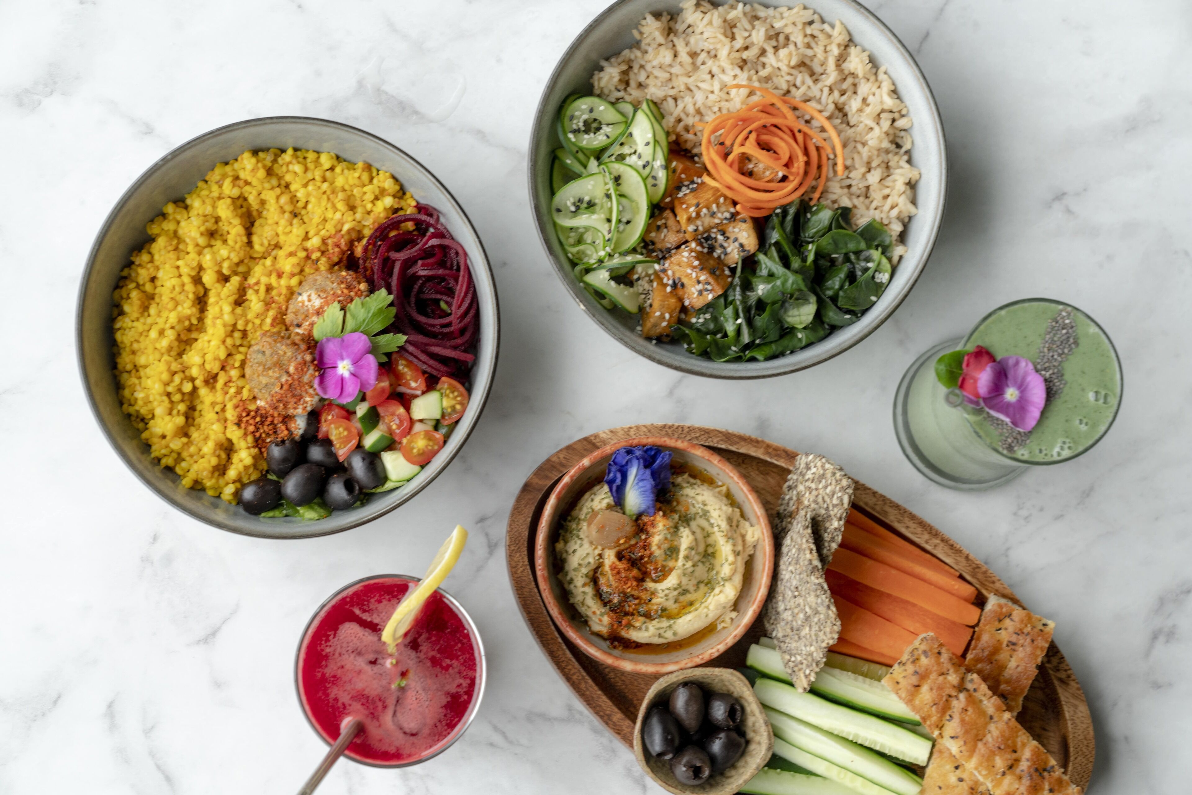 A variety of dishes including a bowl of couscous, a salad with cucumbers and carrots, and a bowl of hummus. There are also some olives, a carrot stick, and a green smoothie.