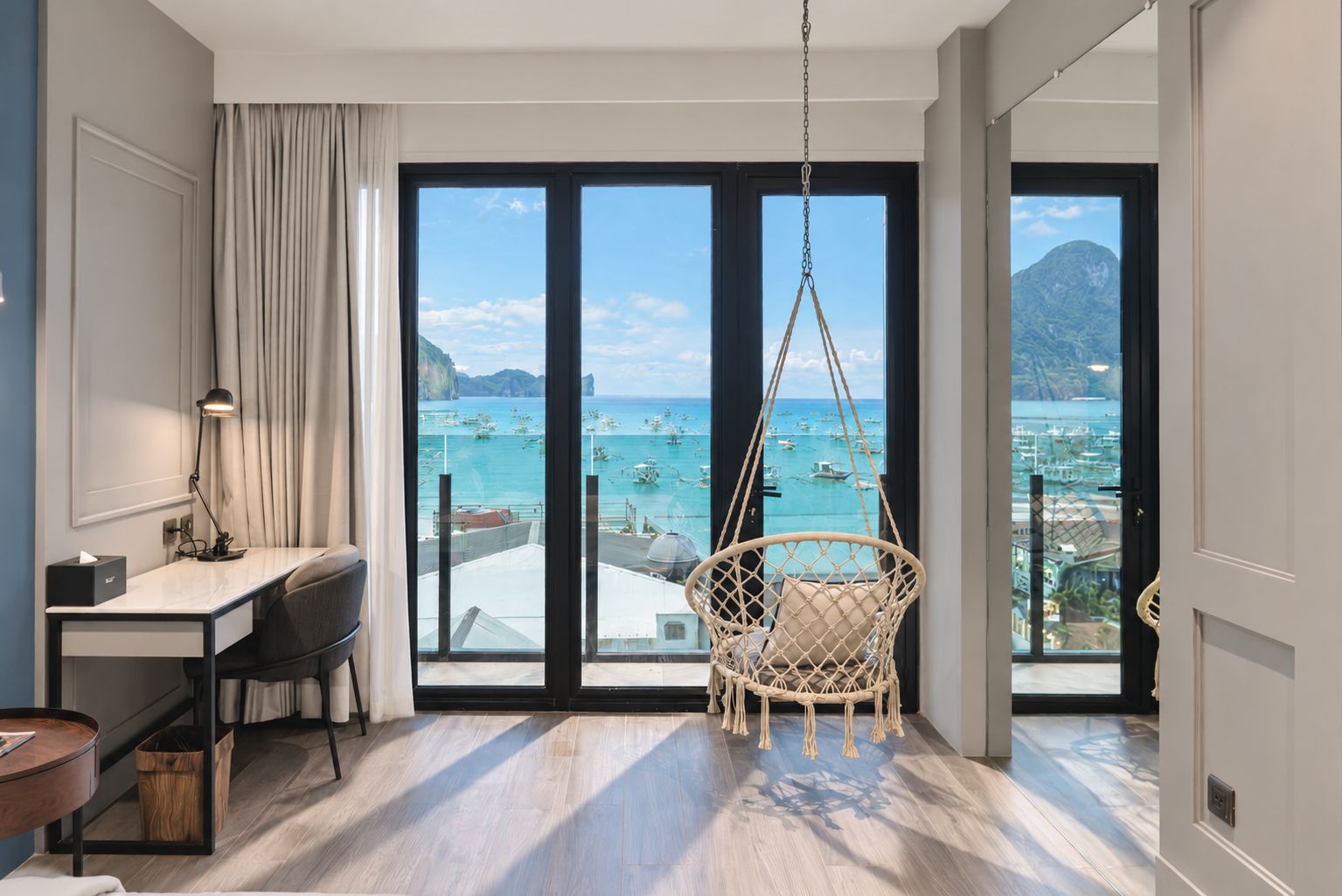 A room with a desk, chair, and a hanging chair with a view of the sea and boats.