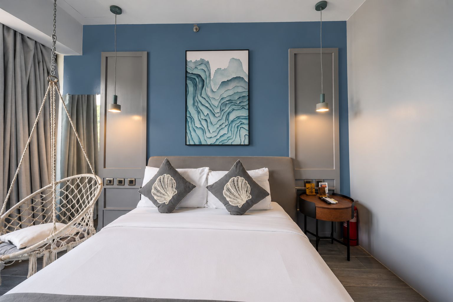 A hotel room with a bed, a hanging chair, a painting, and a bedside table.