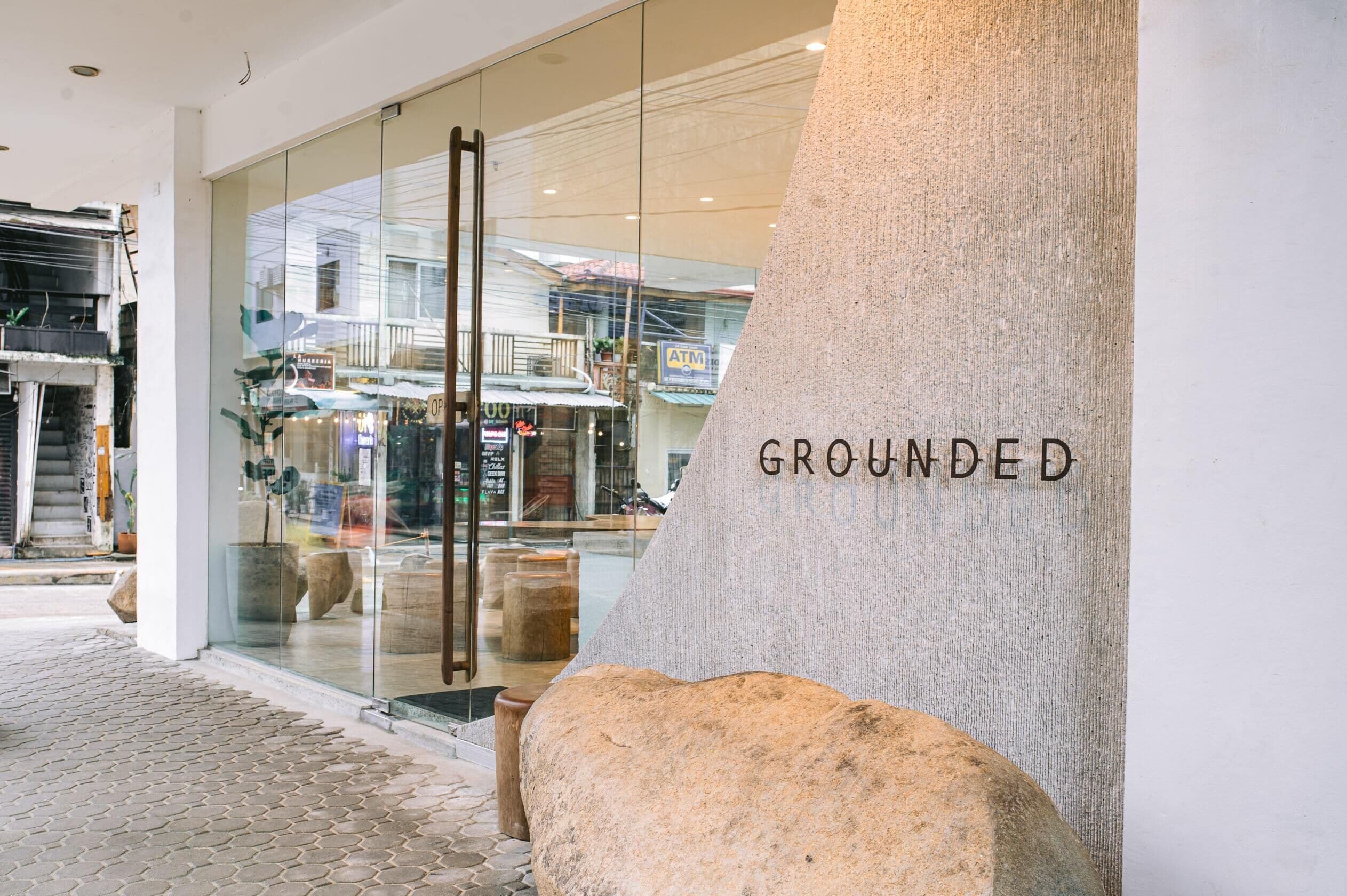 A storefront with a glass facade, a large rock with 'GROUNDED' text, and a cobblestone path.