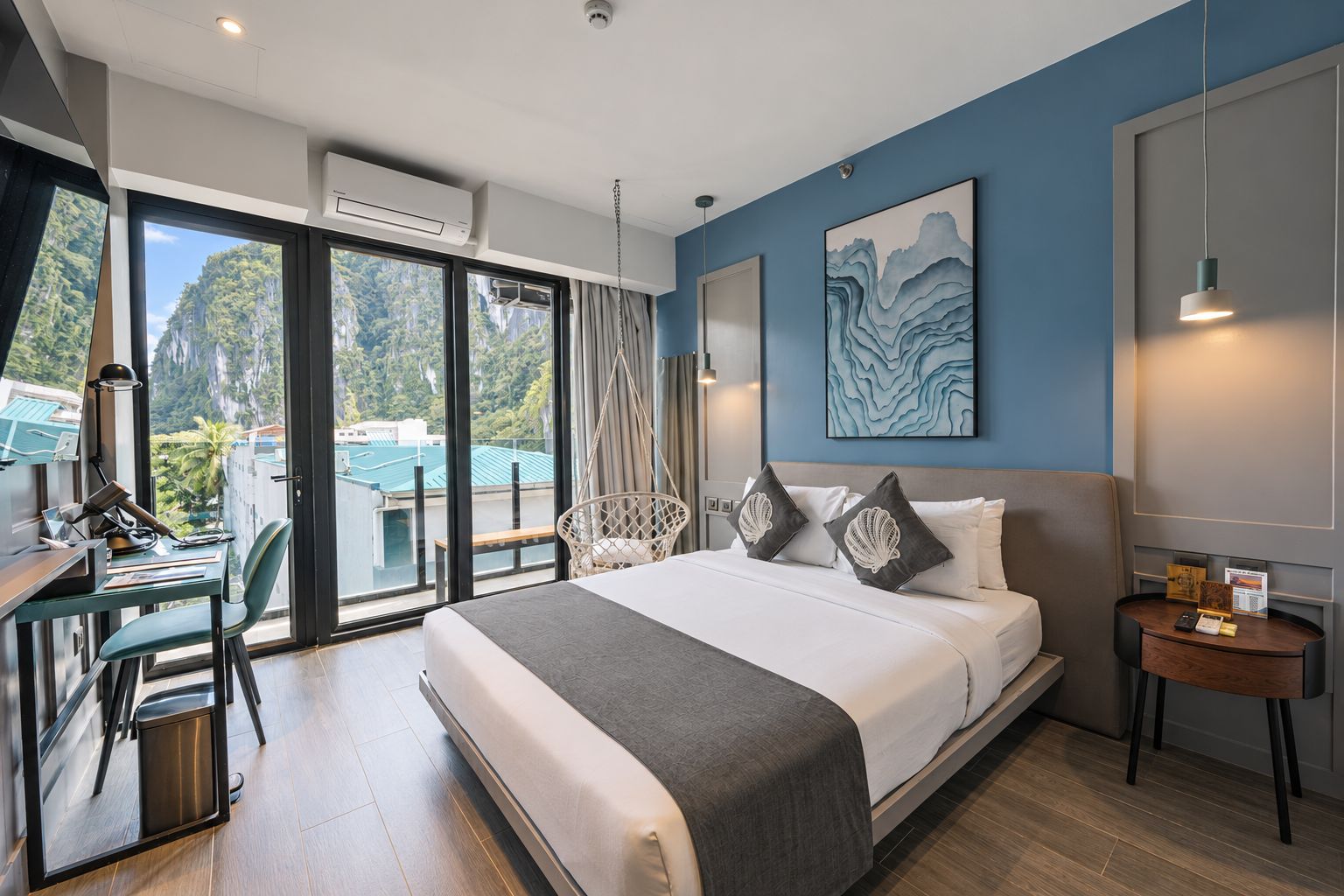 A modern hotel room with a large bed, a desk with a chair, a view of the outdoors, and a painting on the wall.