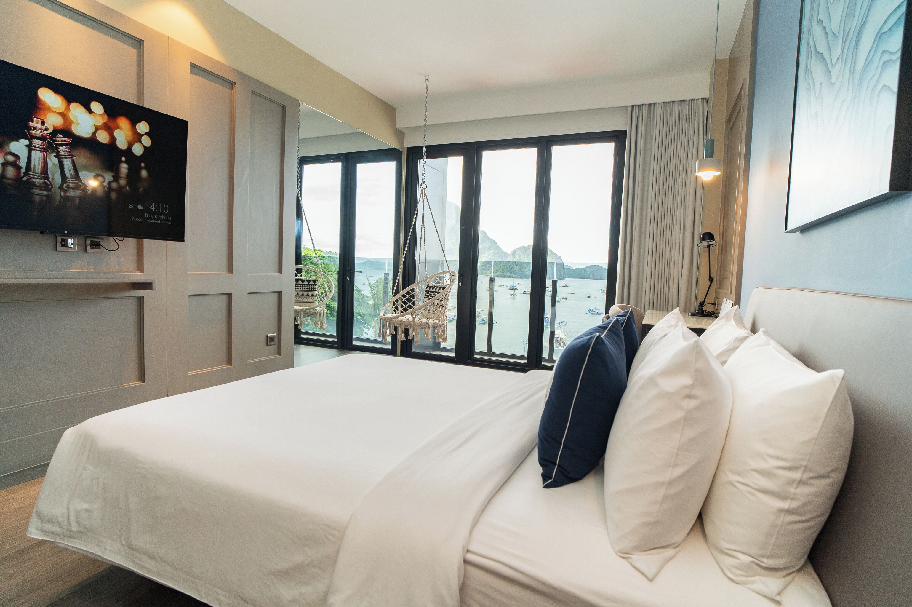 A hotel room with a large bed, a television mounted on the wall, and a view of the sea through large windows.