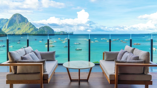 Habitación con dos sofás frente a un ventanal grande con vista al mar turquesa, barcos y un paisaje montañoso.