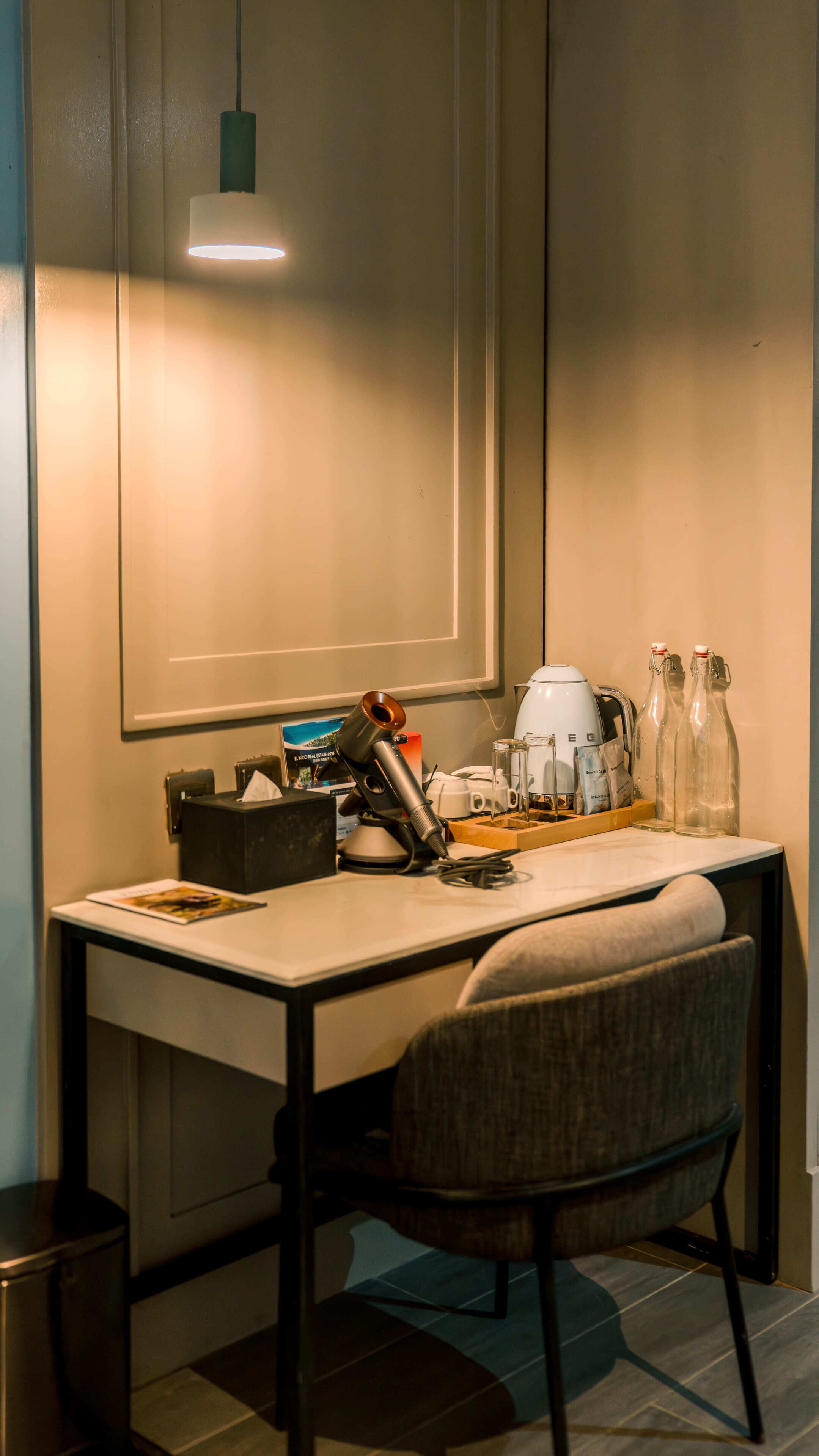 A desk with a chair, a lamp, a kettle, and glass bottles.