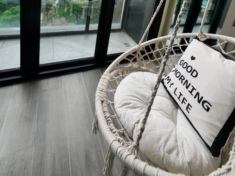 A room with a large window, a hanging chair with a cushion, and a view of greenery and buildings.