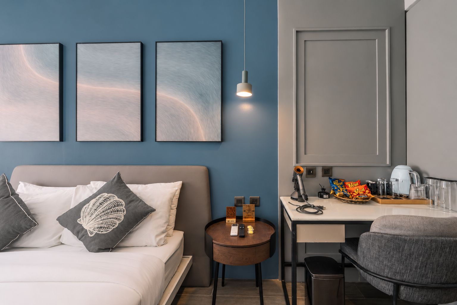 Habitación de hotel con cama, escritorio, silla y una mesita. En la pared, tres cuadros enmarcados, una lámpara y una puerta.