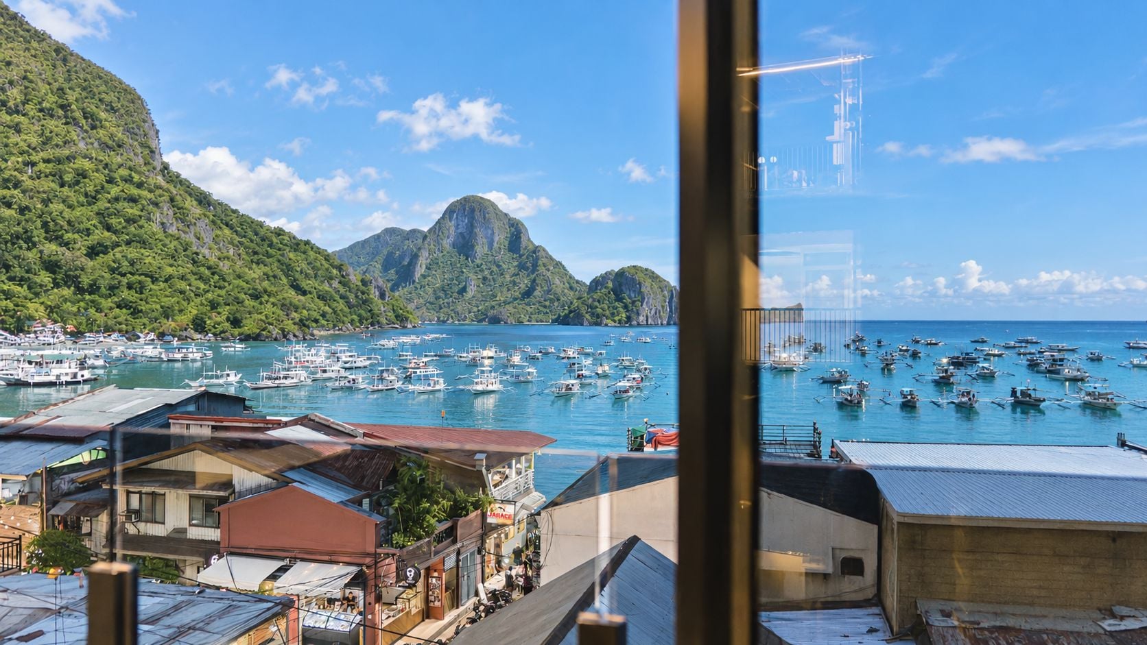 Un pueblo costero con barcos amarrados, montañas al fondo y edificios con señalización visible.