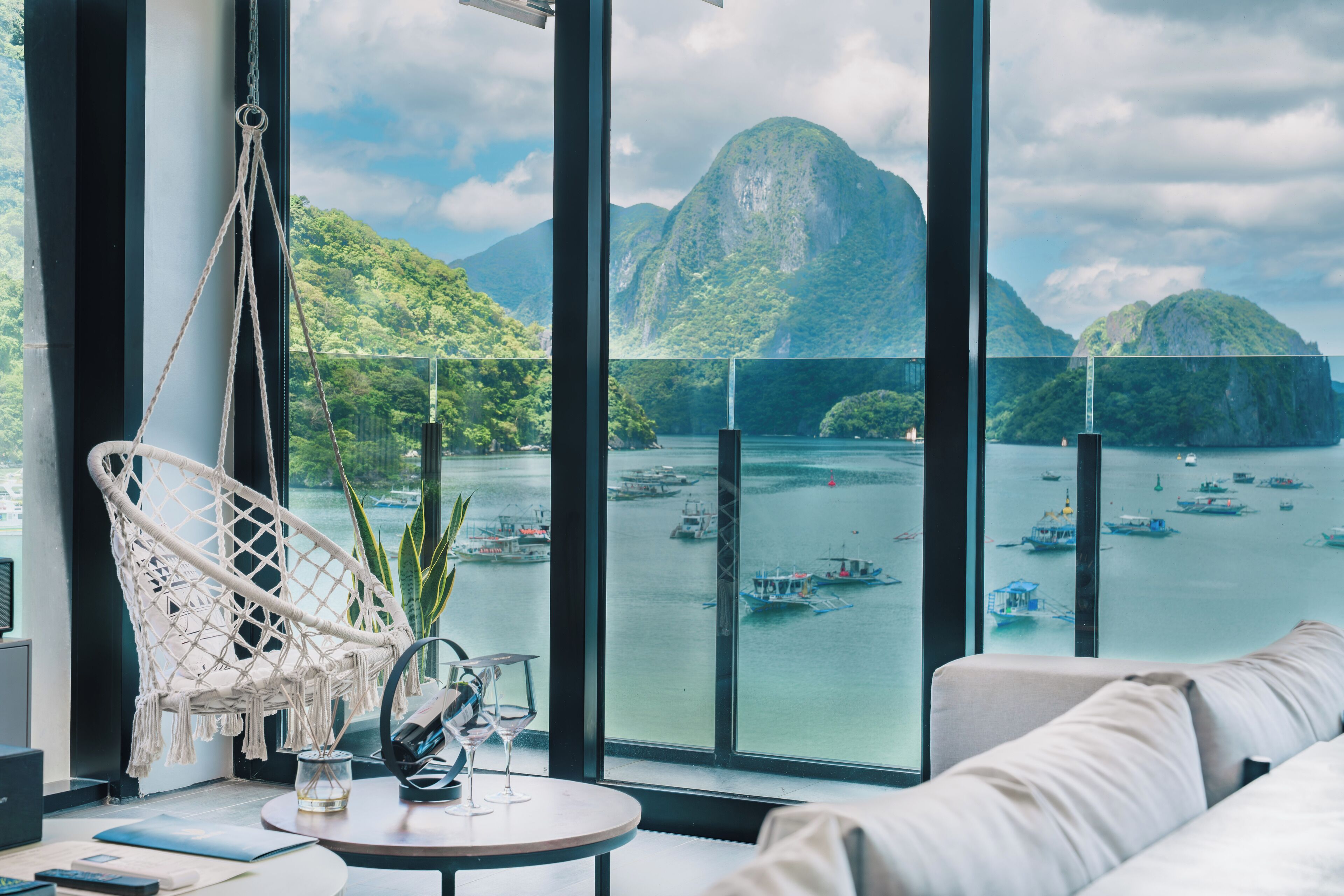 A room with a large window overlooking a bay with boats, a hanging chair, and a round table with a glass and a book.