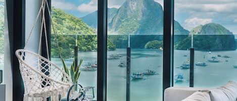 A room with a large window overlooking a bay with boats, a hanging chair, and a round table with a glass and a book.