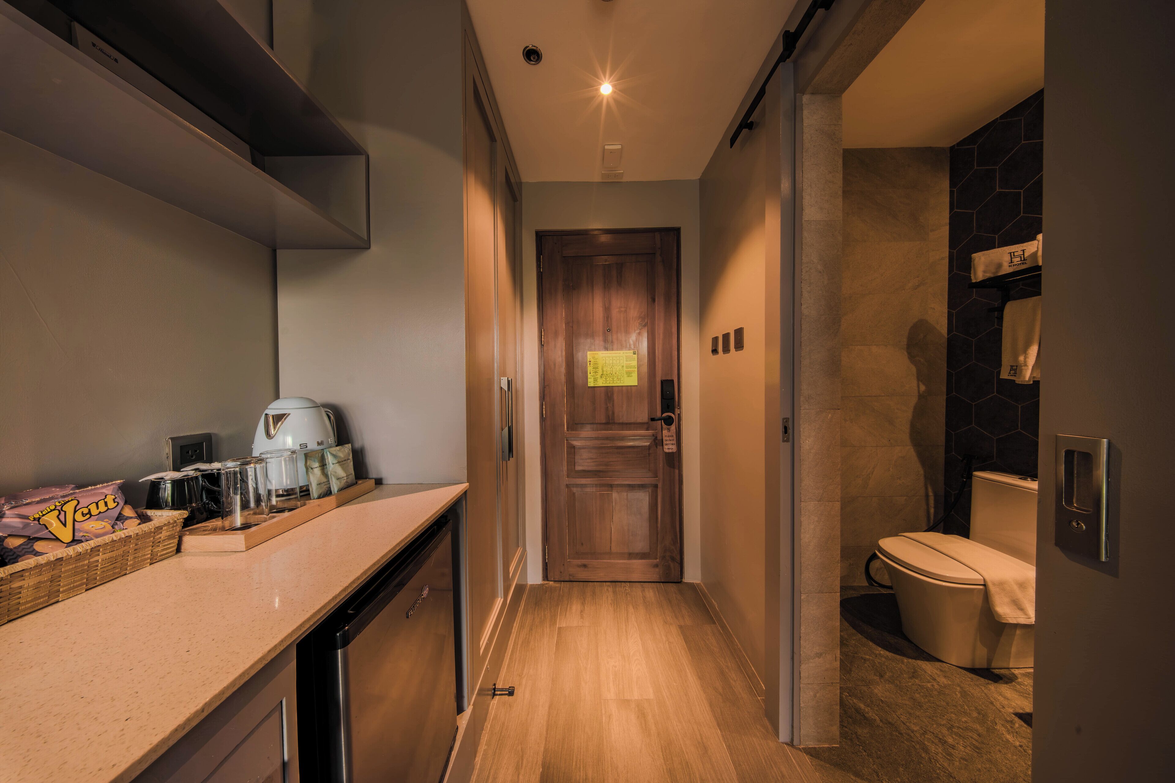A narrow hallway leading to a bathroom with a toilet, a shower, and a sink area with a coffee maker and a basket of snacks.