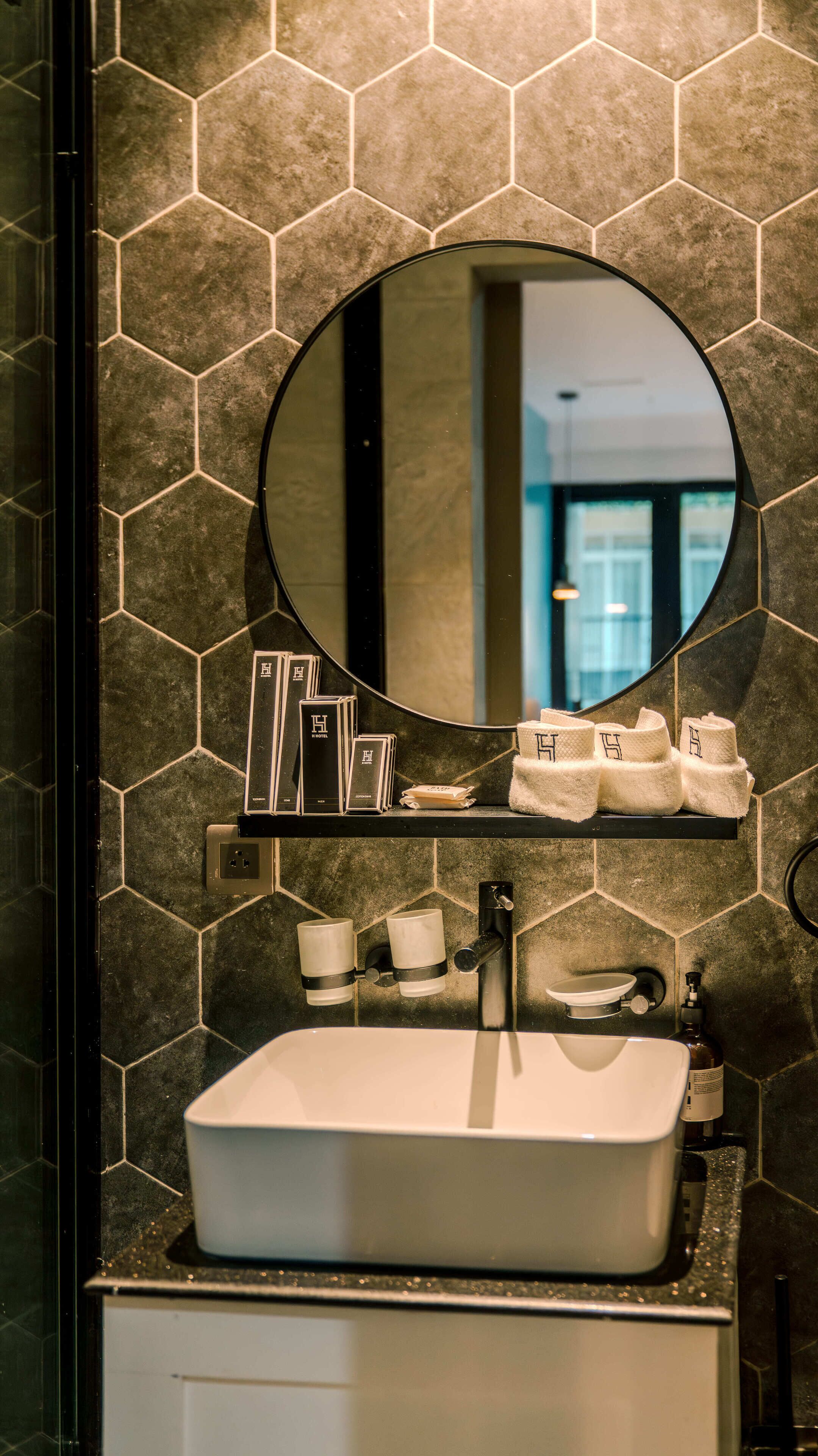 A bathroom with a hexagonal tile backsplash, a round mirror, a white sink, and towel dispensers.