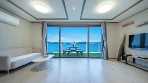A modern living room with a sofa, a coffee table, a TV, and a balcony with a table and chairs. - Yeosu Baraboda Ocean View Pension (Yeosu)