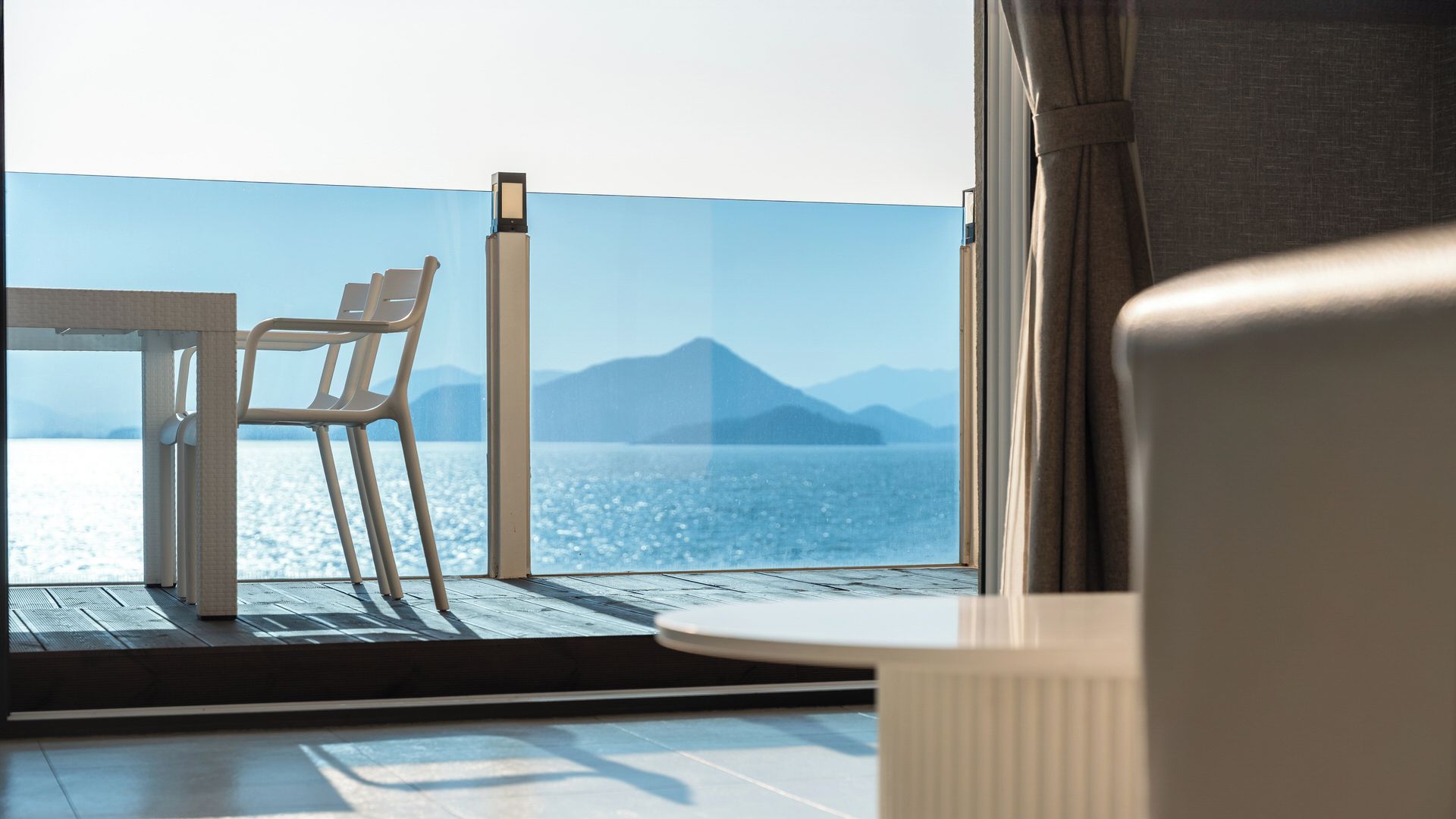 Un balcón con mesa y sillas, con vista a un cuerpo de agua y montañas a lo lejos.