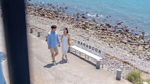 A couple walking along a pebbled beach with a bench labeled 'BARABODA' and a view of the sea.