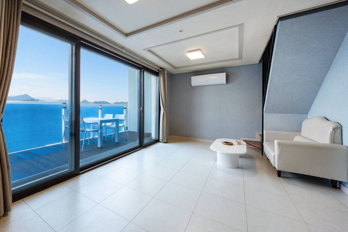 Ett modernt hotellrum med en stor skjutdörr i glas som leder ut till en balkong med utsikt över havet. På balkongen finns en vit soffa, ett litet runt bord och en stol.
