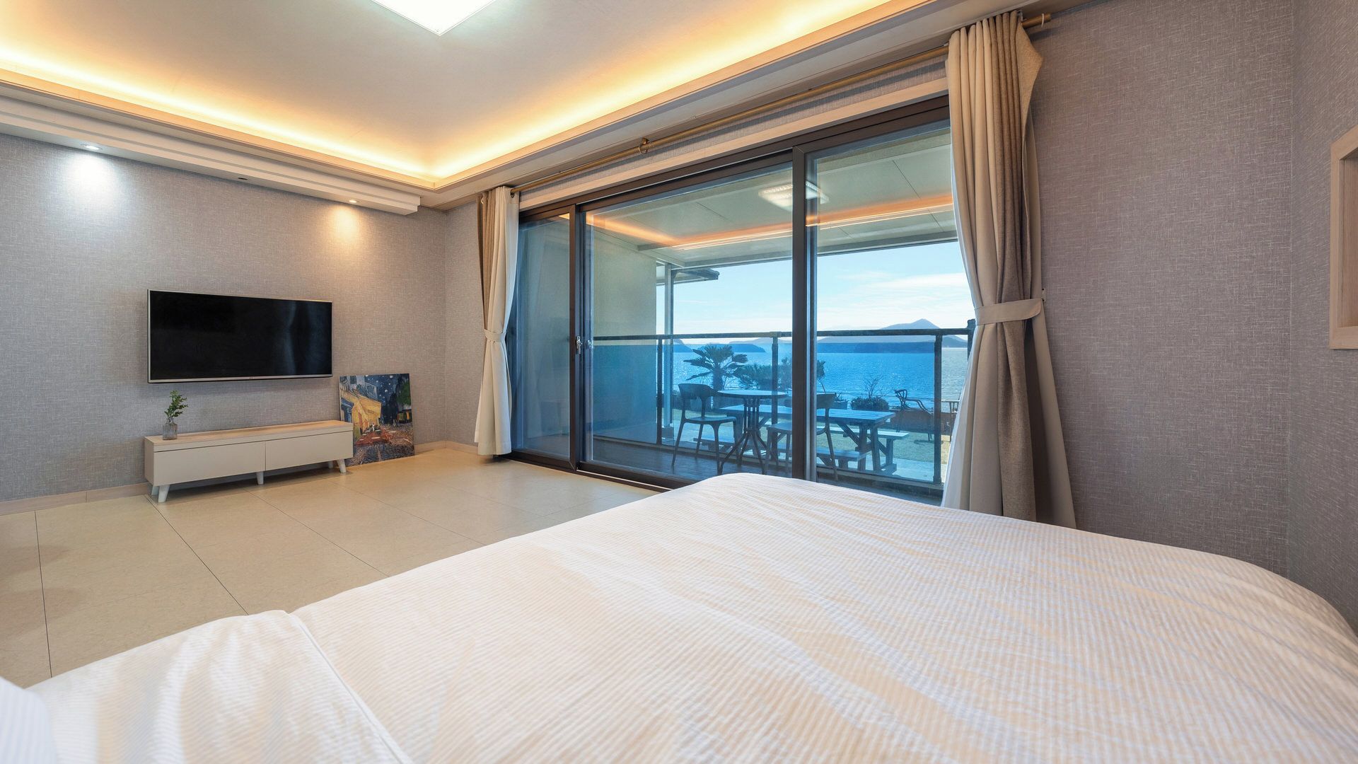 A hotel room with a large bed, a flat-screen TV, a painting, and a balcony with a view of the sea.