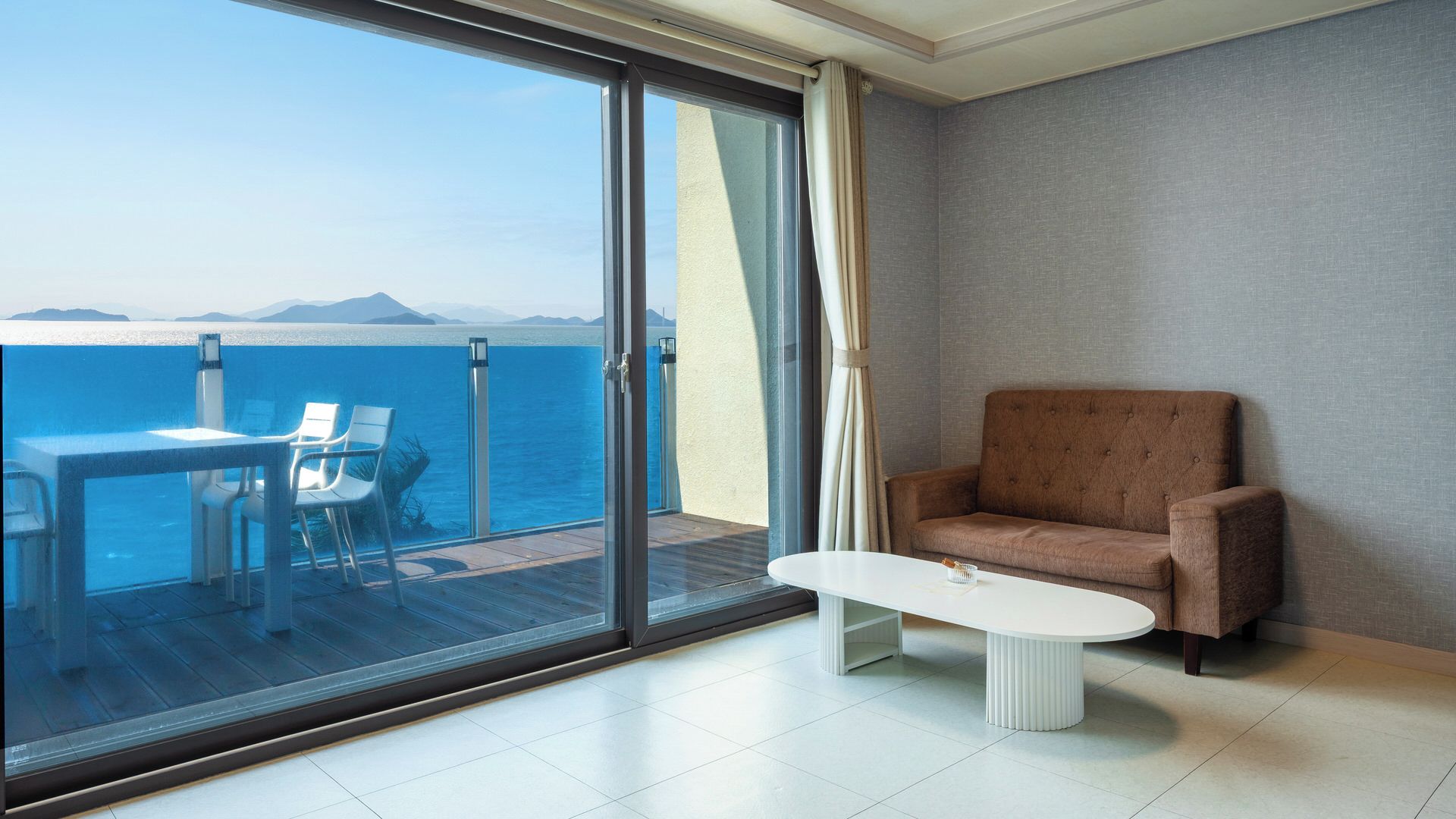 Una habitación moderna con un amplio ventanal corredero que da a un balcón con vistas al mar y a las montañas.
