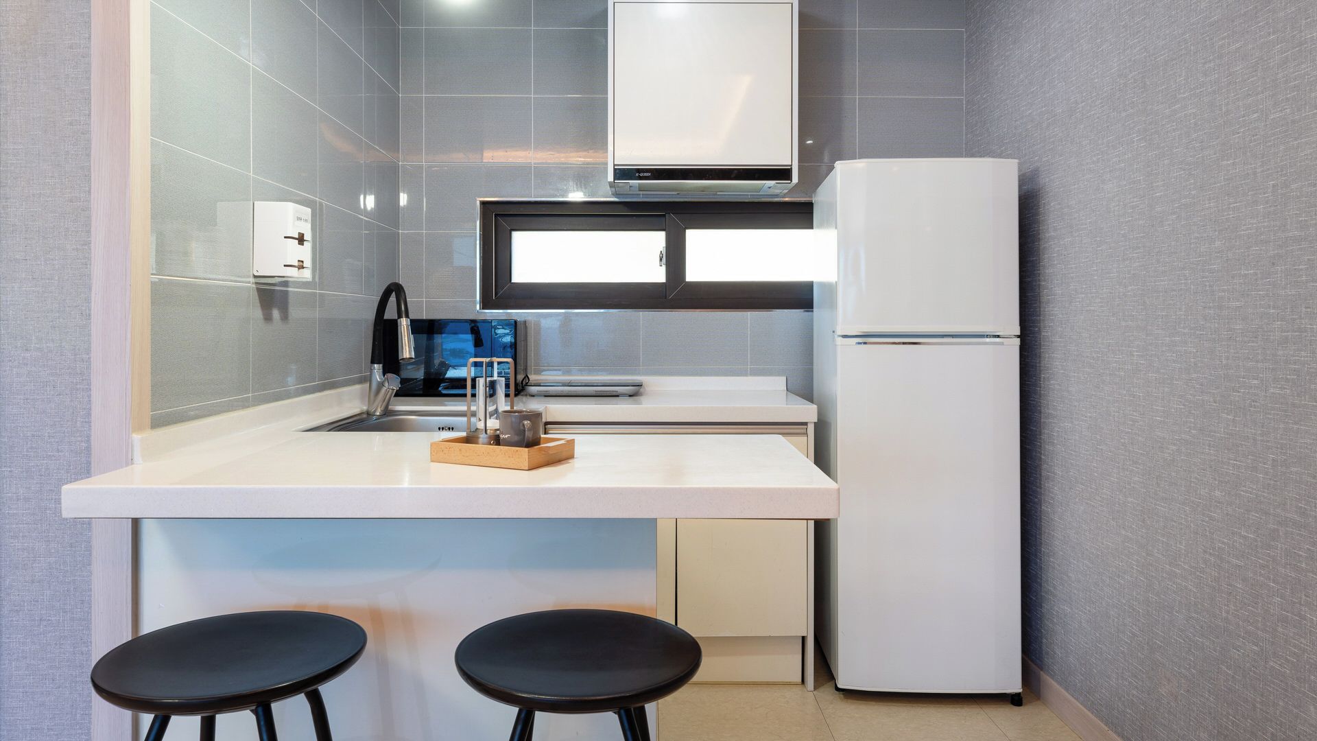 A compact kitchen with a white refrigerator, a sink, and a window above the counter. There are two black stools at the counter.