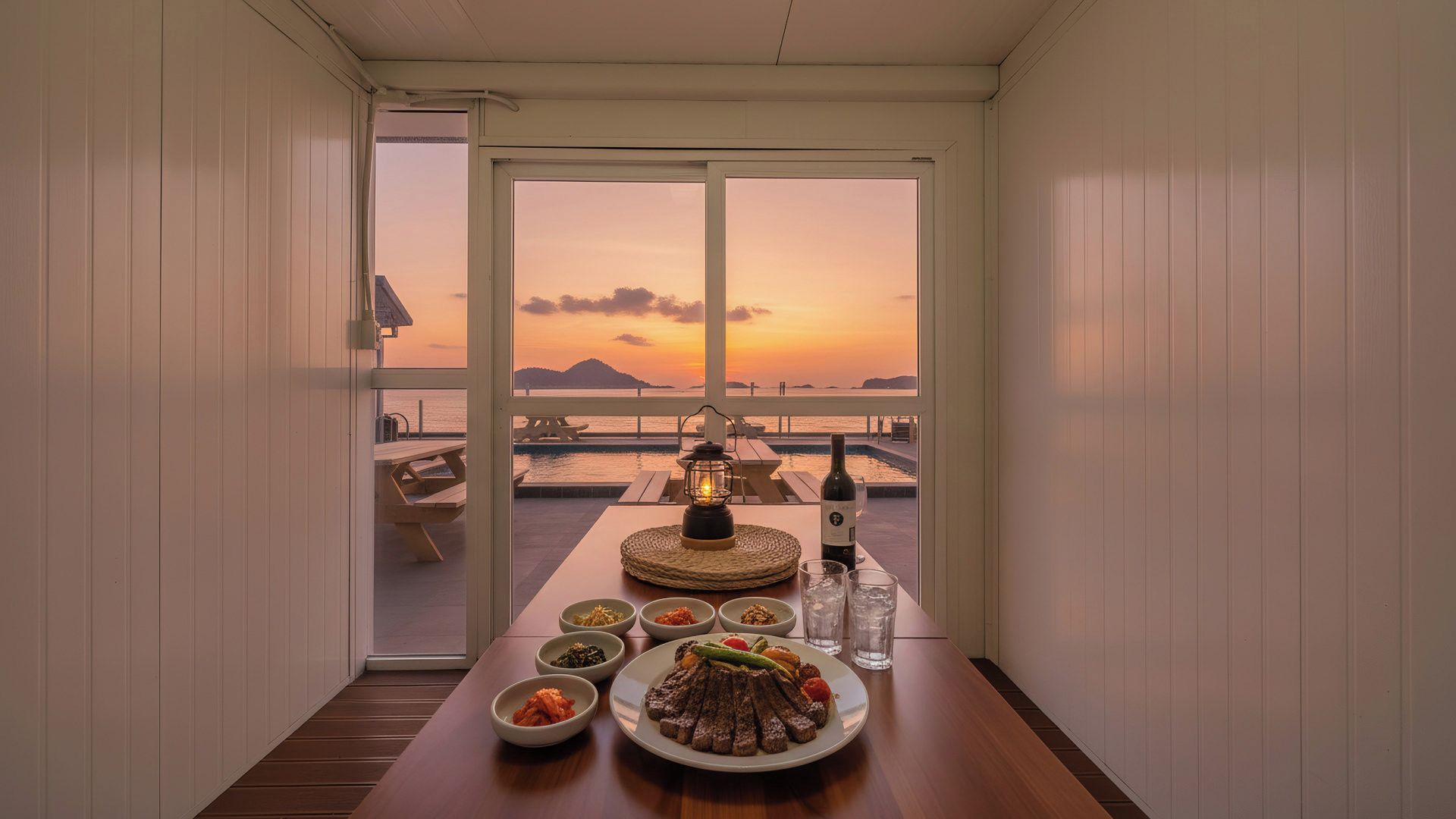 A dining area with a table set for a meal, a bottle of wine, and a view of the sunset over the water.
