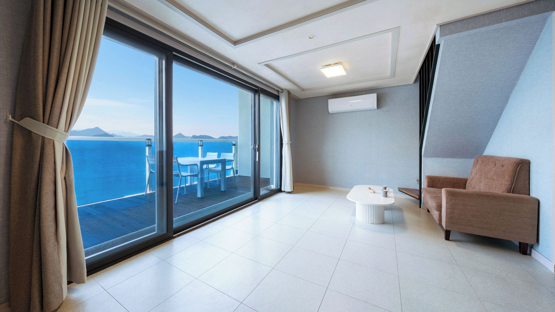 A modern hotel room with a large sliding glass door leading to a balcony with a view of the sea and mountains.
